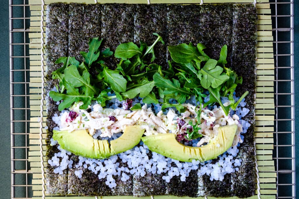 shirataki rice, avocado, chicken salad and arugula spread on a nori sheet, placed on a bamboo mat before rolling