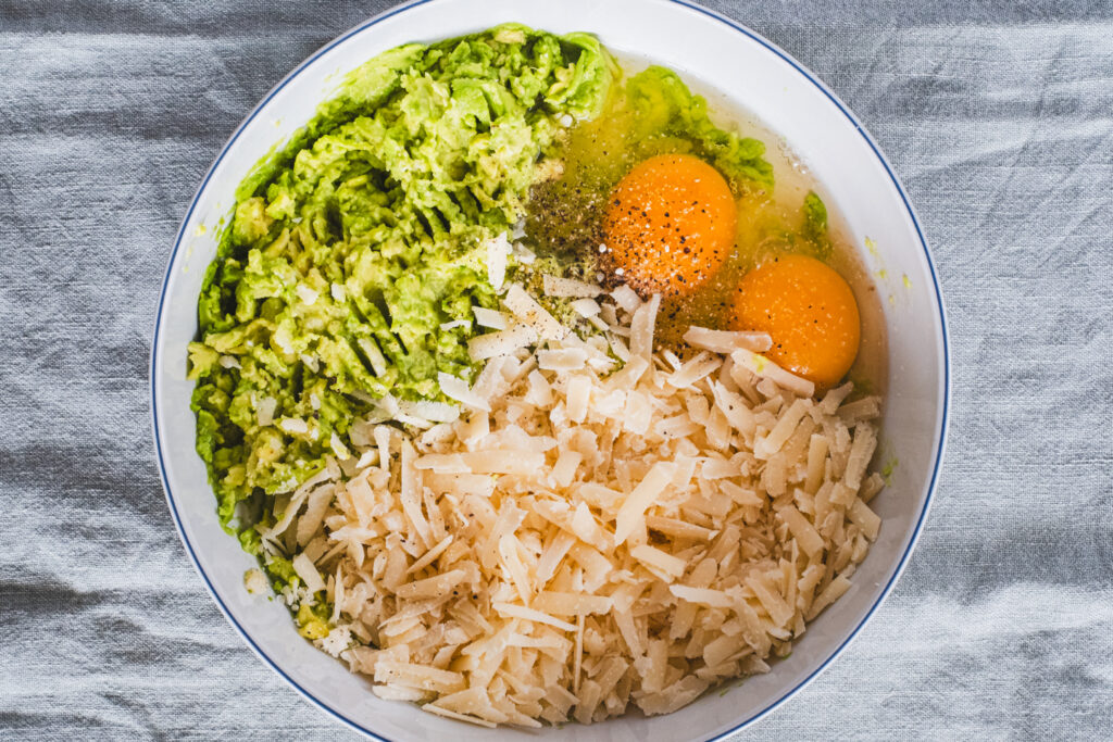 A mixing bowl containing mashed avocados, eggs, grated cheese, and seasonings.