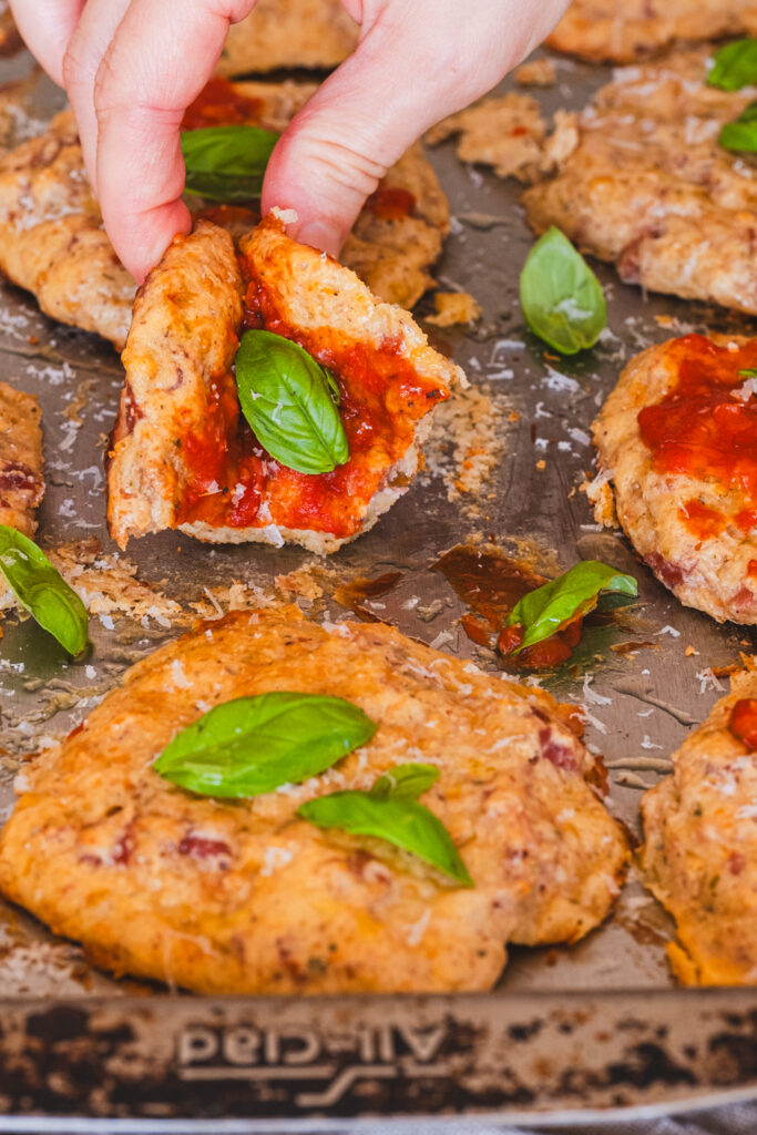 hand picking up a cottage cheese pizza bite on a baking sheet, topped with ketchup and basil leaves