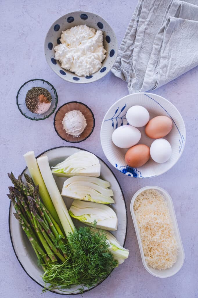 A display of fresh ingredients including leeks, fennel, and asparagus, arranged together for a crustless vegetable quiche.