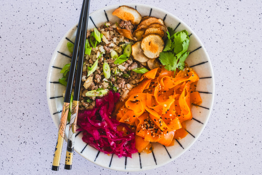 A colorful bowl featuring seasoned ground beef and cauliflower rice, accompanied by tangy homemade sauerkraut and a fresh carrot ribbon salad.