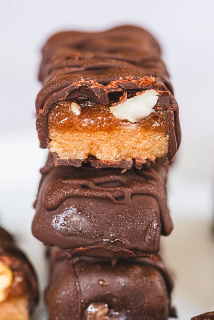 A stack of healthy Snickers bars, with the top bar cut in half to clearly display the layered base, visible nuts, and caramel.