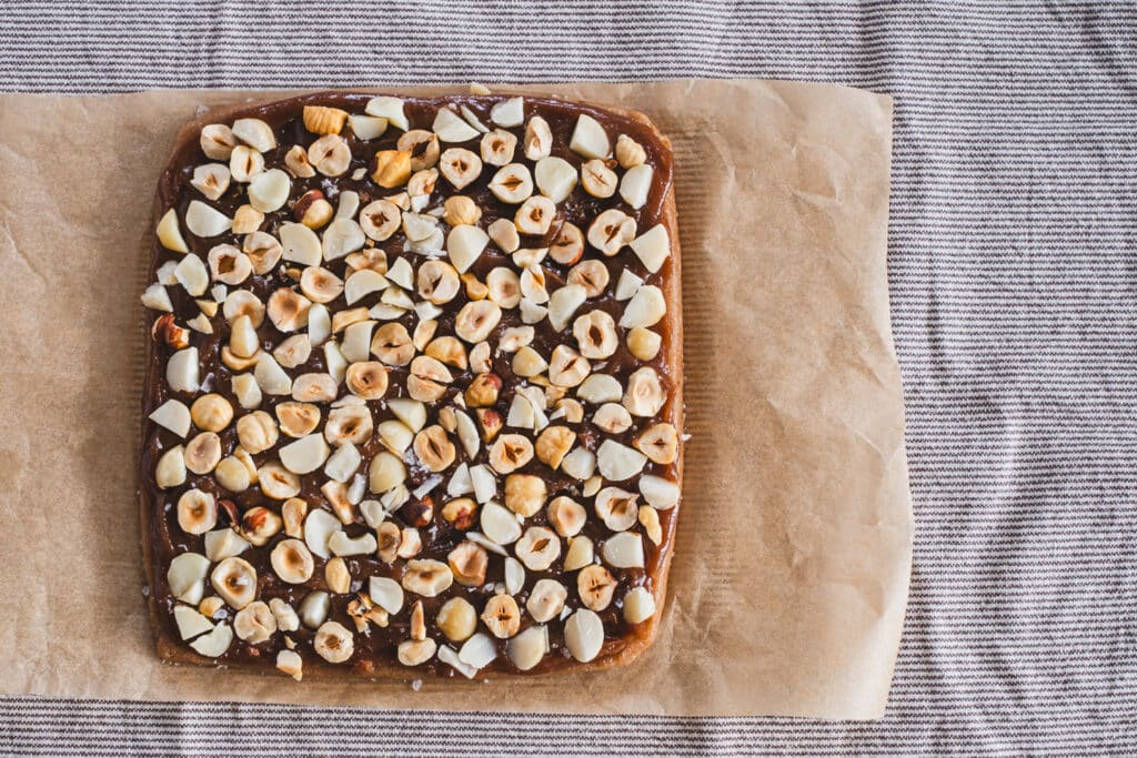 A layer of chopped nuts is firmly pressed into the glossy caramel surface atop the base layer.