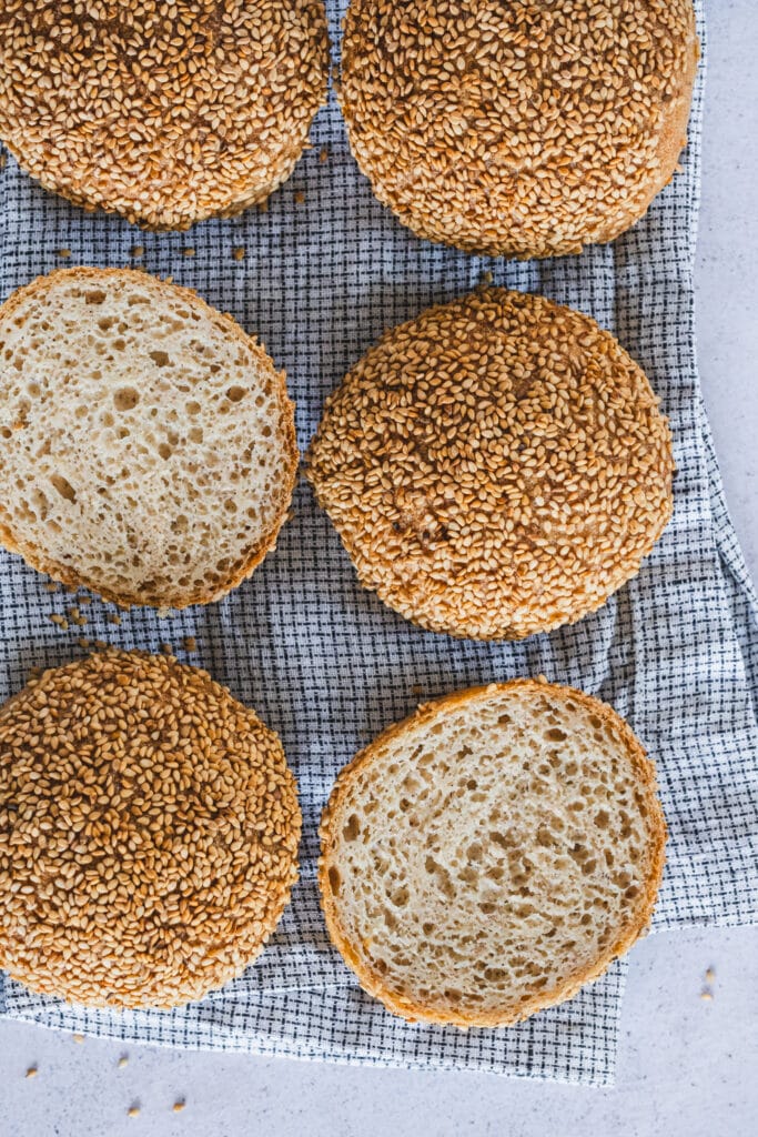 Sesame-crusted gluten-free sourdough buns on a kitchen towel. One has been sliced in half to show the texture.