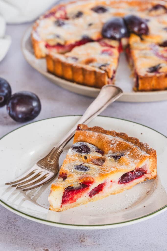 Gluten free plum custard tart slice on a plate, served with a fork, with the full tart in the background