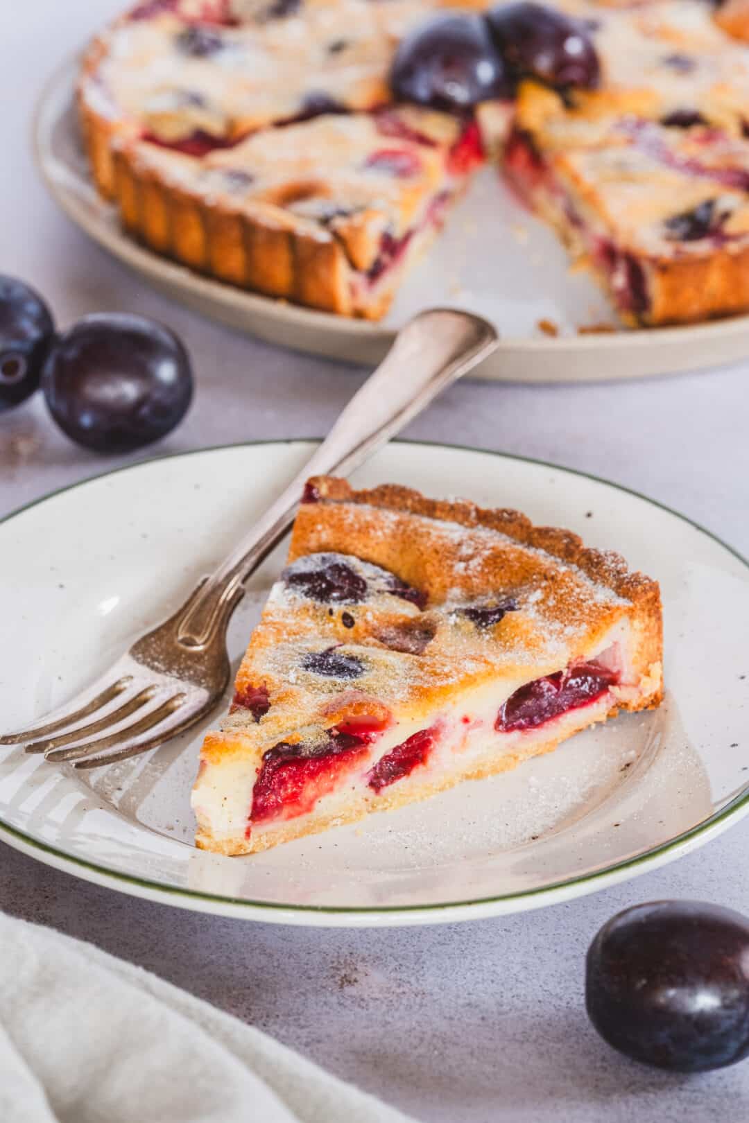 Gluten free plum custard tart slice, served on a plate, a fork, and a full tart in the background