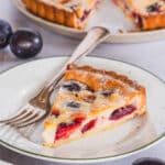 Gluten free plum custard tart slice, served on a plate, a fork, and a full tart in the background
