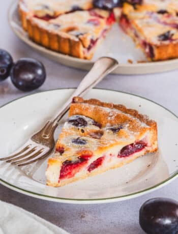 Gluten free plum custard tart slice, served on a plate, a fork, and a full tart in the background
