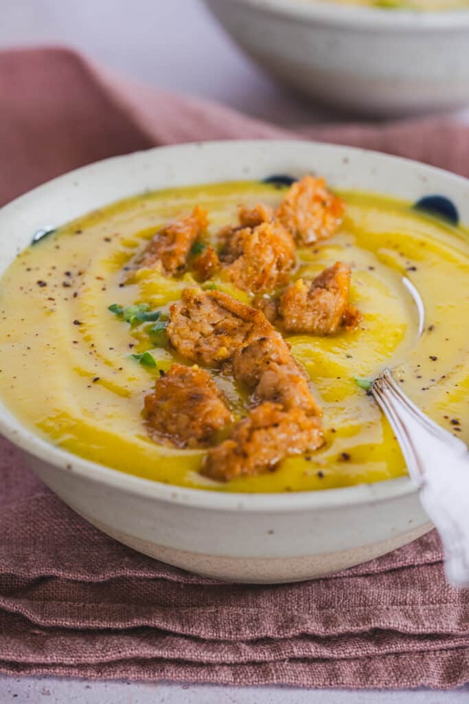 A bowl with yellow creamy soup topped with homemade Italian chicken sausage bites, microgreens, and a spoon for serving